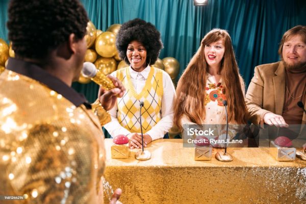 A fun depiction of a competitive TV game show, stylized in late 1970s or early 1980s fashion.  The host, an African American man in a stunning gold blazer, asks the contestants quiz trivia questions to see who will win the grand prize!  The participants hold their hands over their buzzers to signal they have the answer.